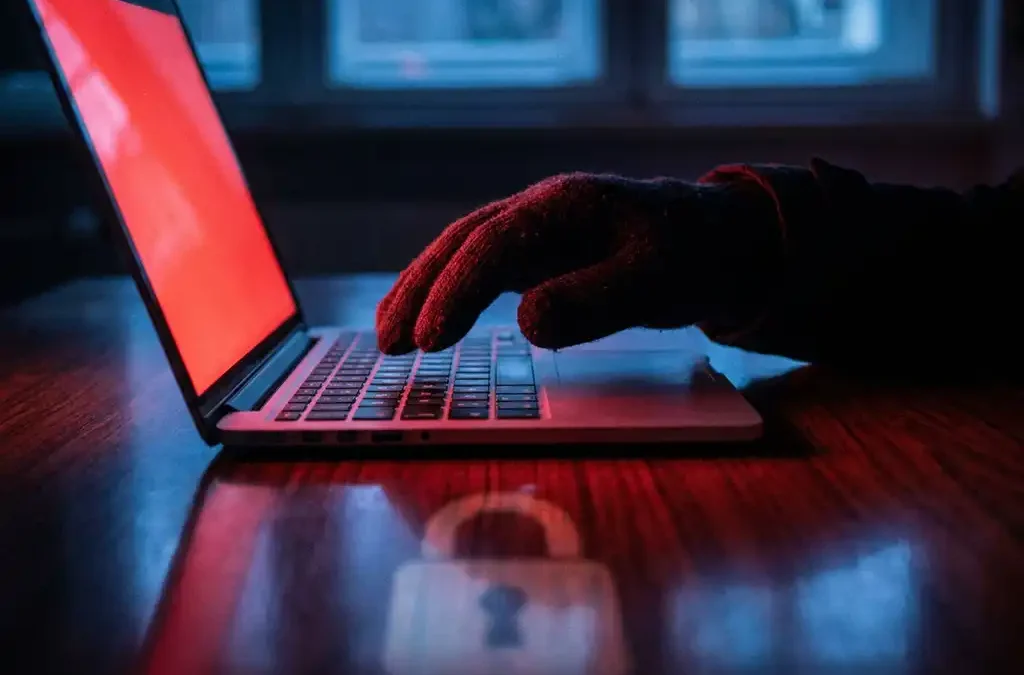 Gehandschoende hand reikt naar laptoptoetsenbord, rood schermgloed en slotsymbool weerspiegeld op donker bureauoppervlak.